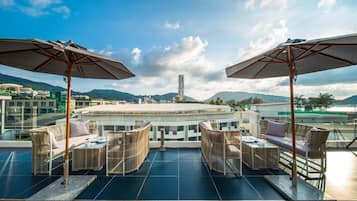 A rooftop terrace with wicker furniture, umbrellas, and a view of a cityscape and mountains.