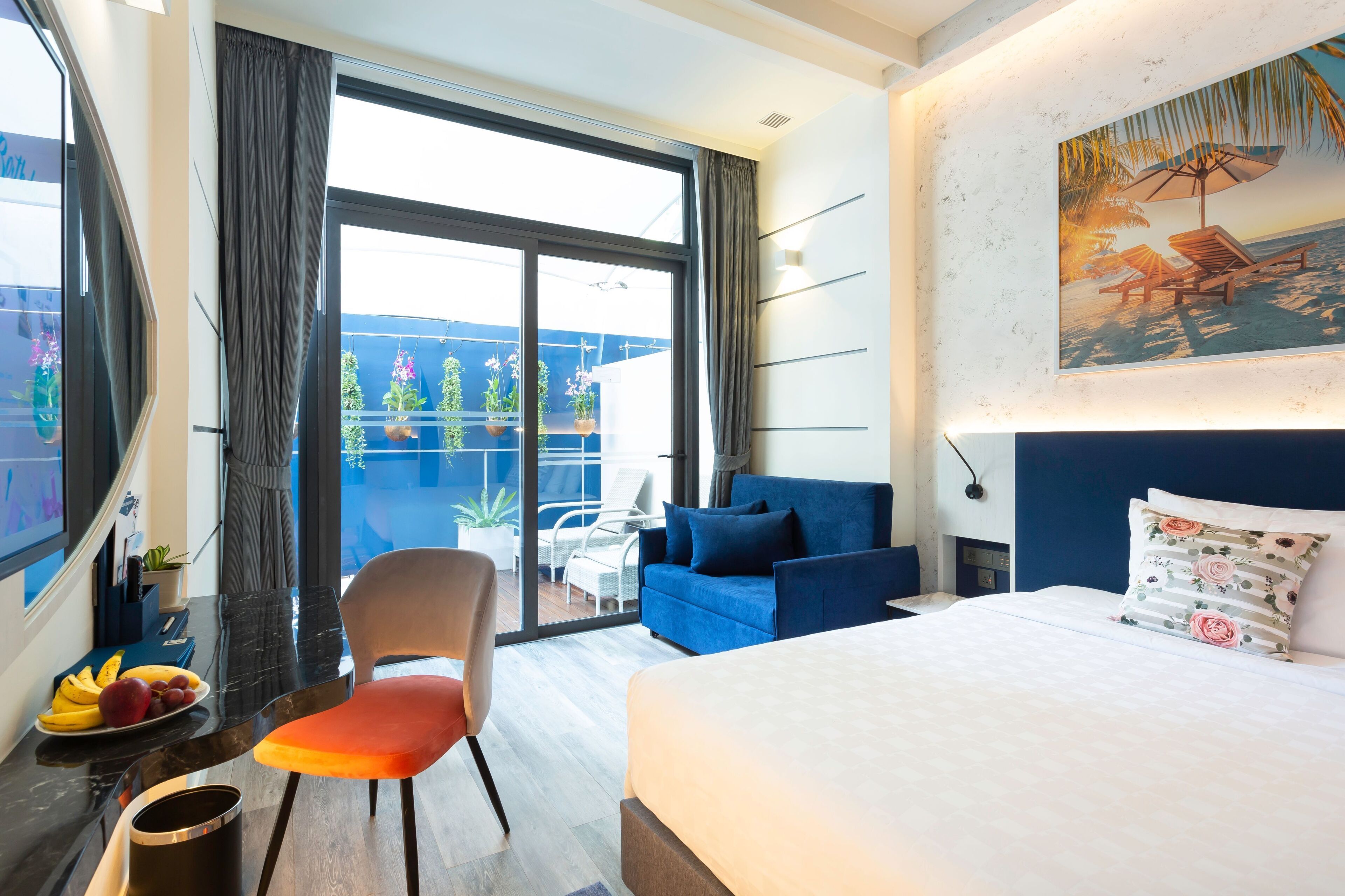 A modern hotel room with a large bed, a blue sofa, a dining table with fruit, and a balcony with plants and chairs.