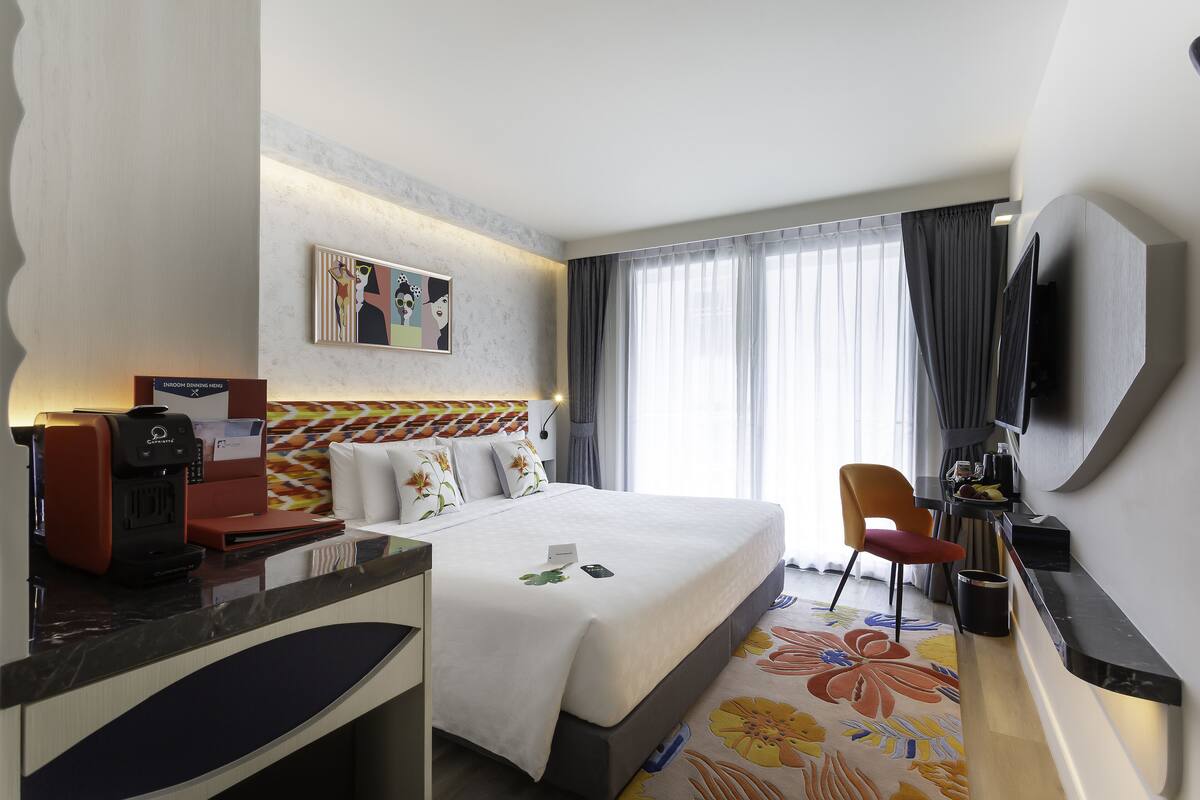 Habitación de hotel con cama, escritorio con cafetera, televisor montado en la pared y una ventana grande con cortinas.