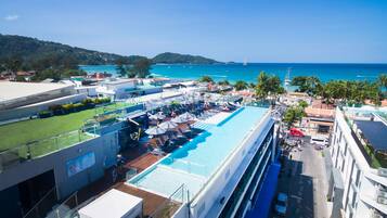 A rooftop pool area with multiple lounge chairs and umbrellas, overlooking a coastal landscape with clear blue waters and distant hills.