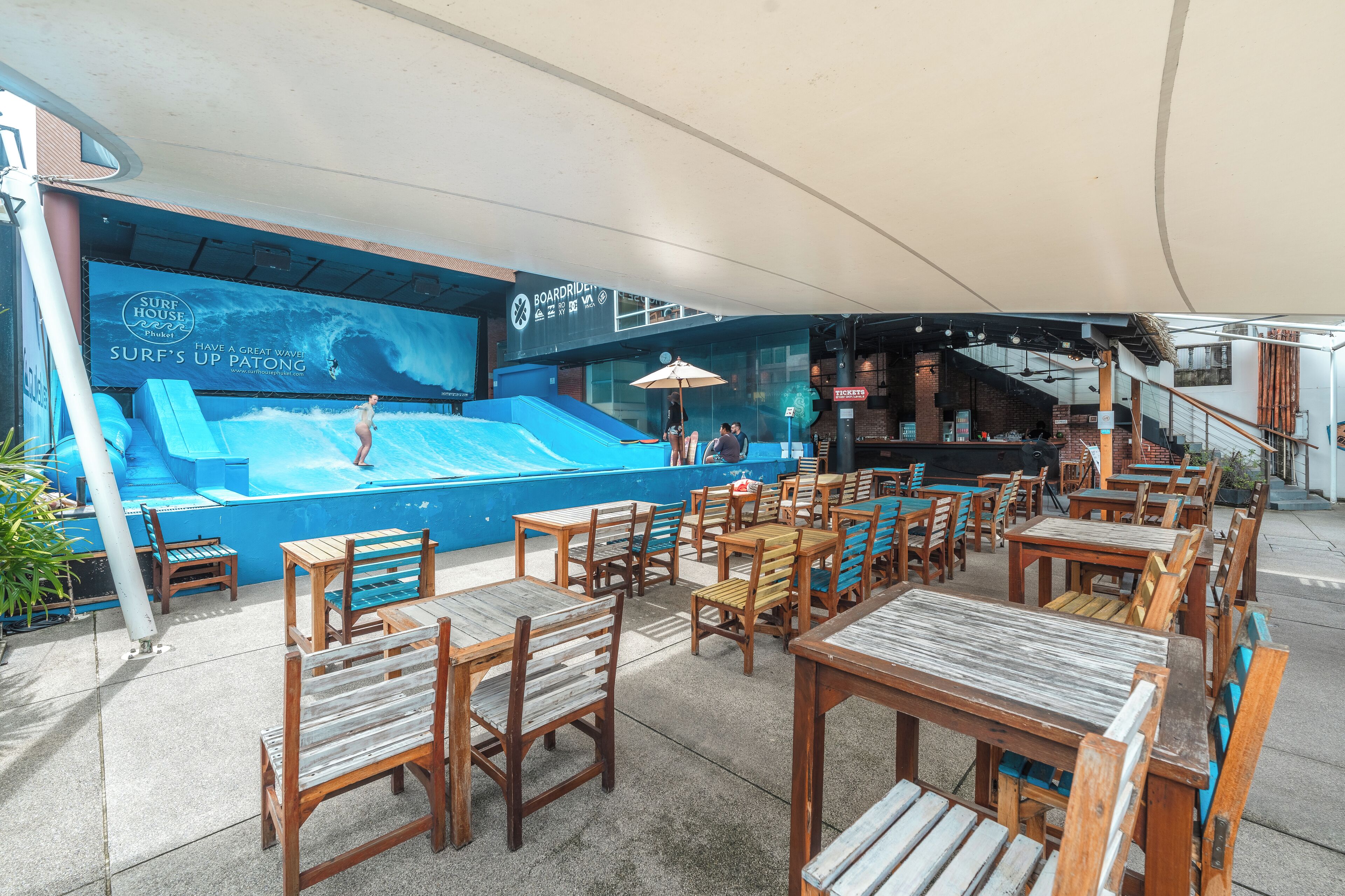 An outdoor seating area with wooden tables and chairs, a pool with a wave design, and a large screen displaying a surf advertisement.