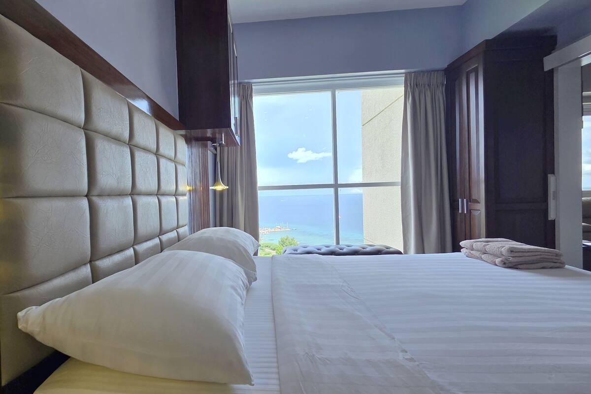 A hotel room with a large bed, a view of the sea, and a window with curtains.