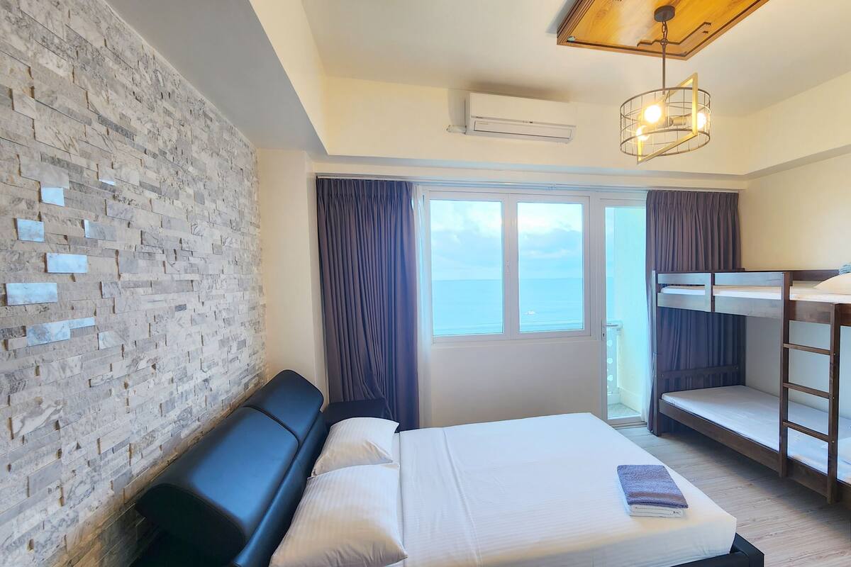 A hotel room with a bed, a bench, a wall with a brick pattern, a window, and a ceiling light fixture.