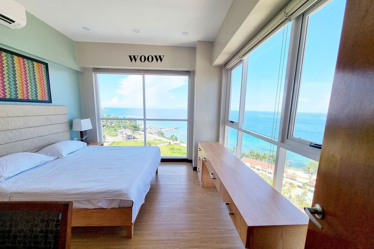 A bedroom with a large bed, a bench, a desk, and a view of the ocean.