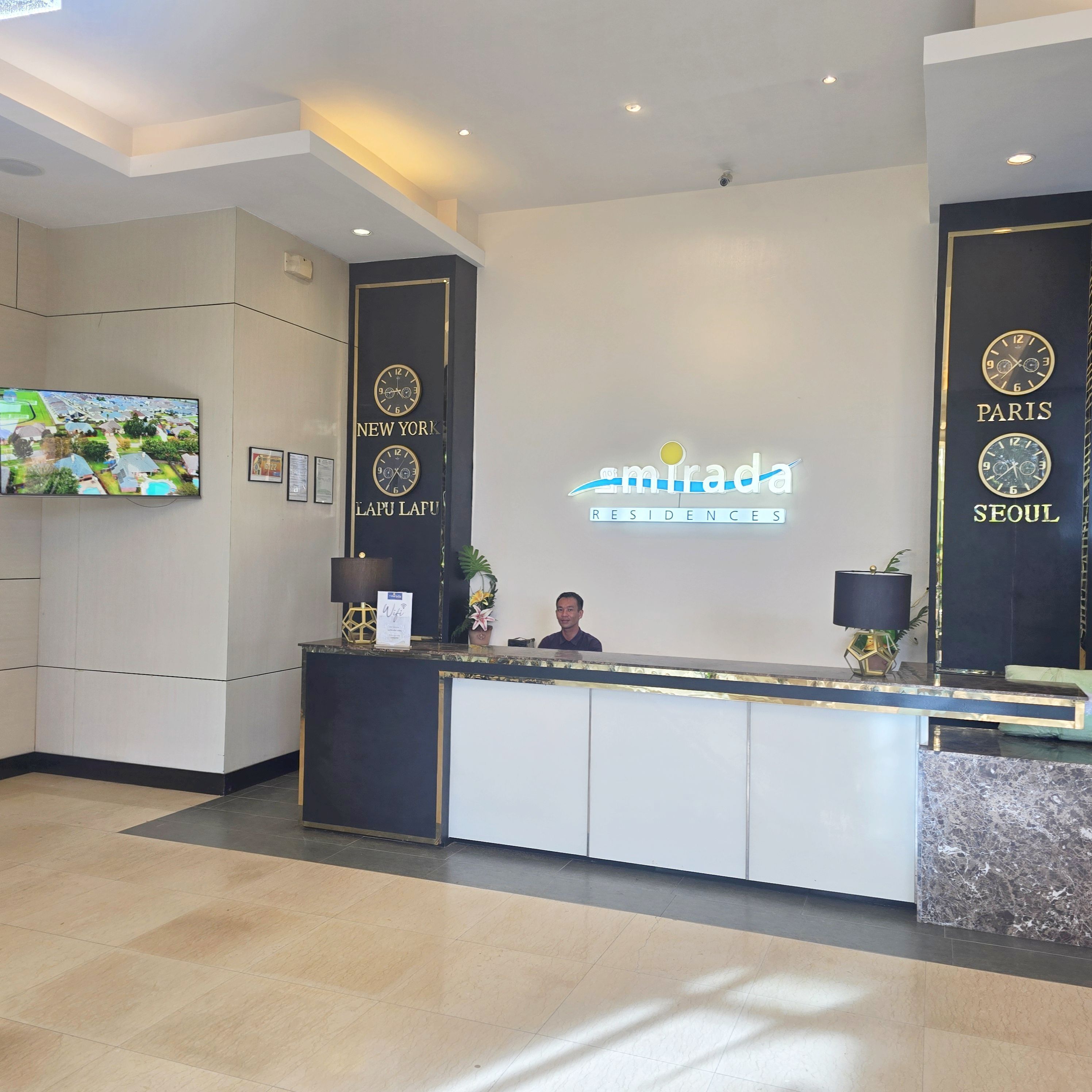 A hotel reception desk with a digital clock displaying 'New York' and 'Paris' on the wall, a flat-screen TV mounted on the wall, and a receptionist behind the desk.