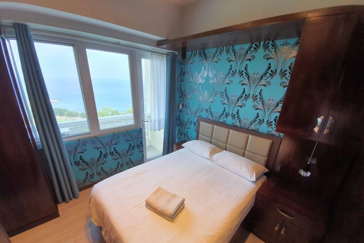 A bedroom with a bed, a window with curtains, and a view of the ocean.