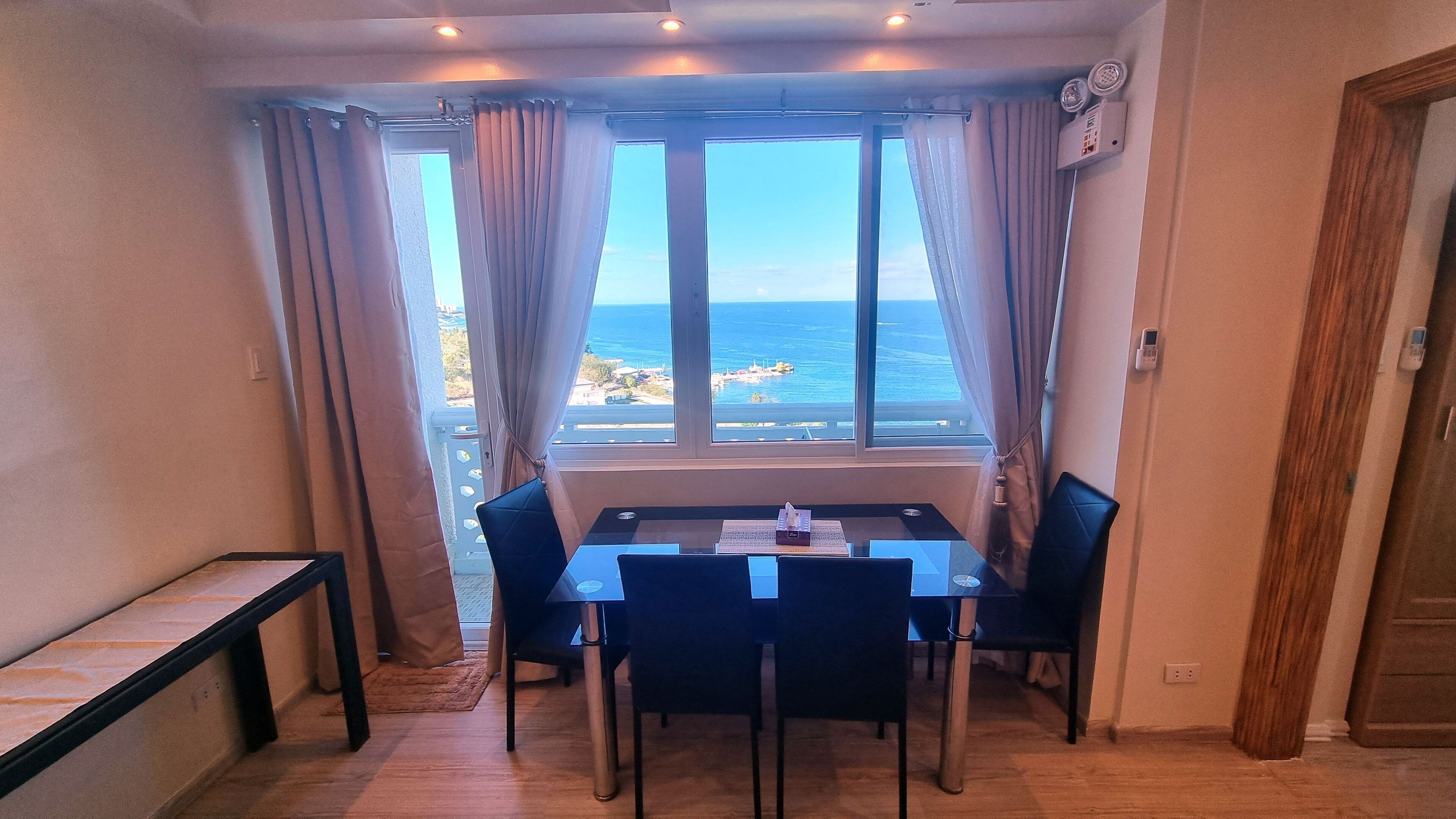 A room with a table and chairs, a window with curtains, and a view of the sea.