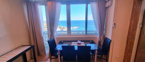 A room with a table and chairs, a window with curtains, and a view of the sea.