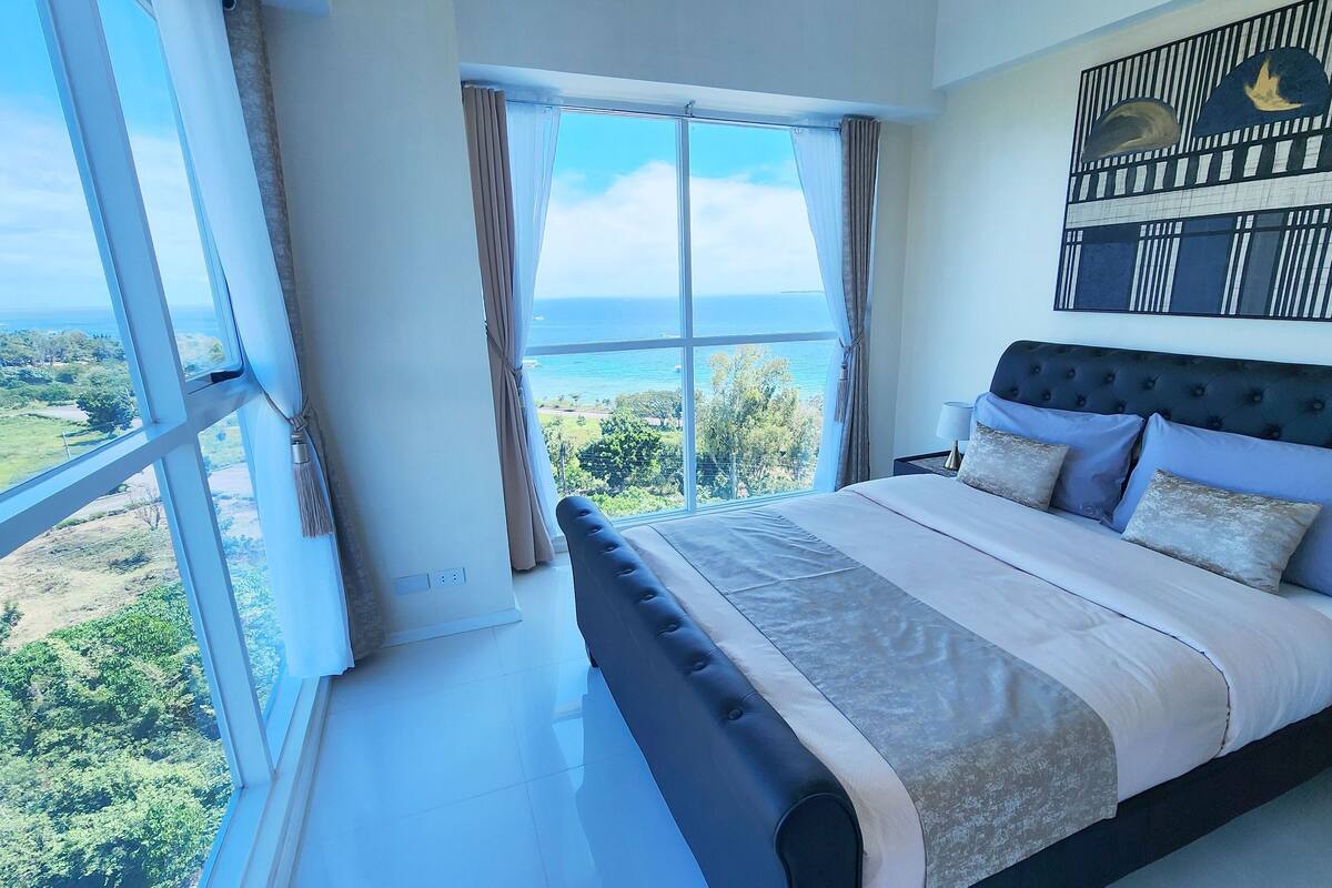 A bedroom with a large window, a bed with a headboard, and a nightstand.