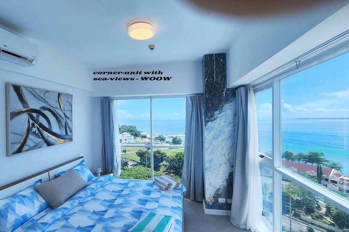 A bedroom with a large window, a bed with a blue and white patterned bedspread, a ceiling light, and a wall-mounted air conditioner.