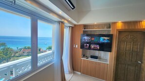 Una habitación con un amplio ventanal con vistas al mar, un televisor instalado mostrando diferentes programas, y una puerta de madera.