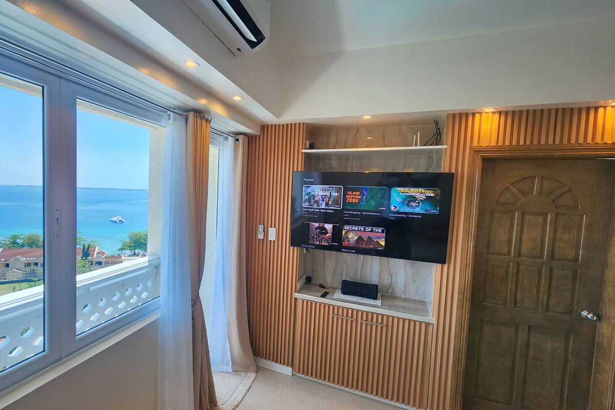 A room with a large window overlooking the sea, a mounted TV displaying various shows, and a wooden door.