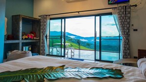 A hotel room with a large window offering a mountain view, a flat-screen TV mounted on the wall, and a bed with a leaf-patterned pillow. - Phu Sandao Resort (Khao Kho)