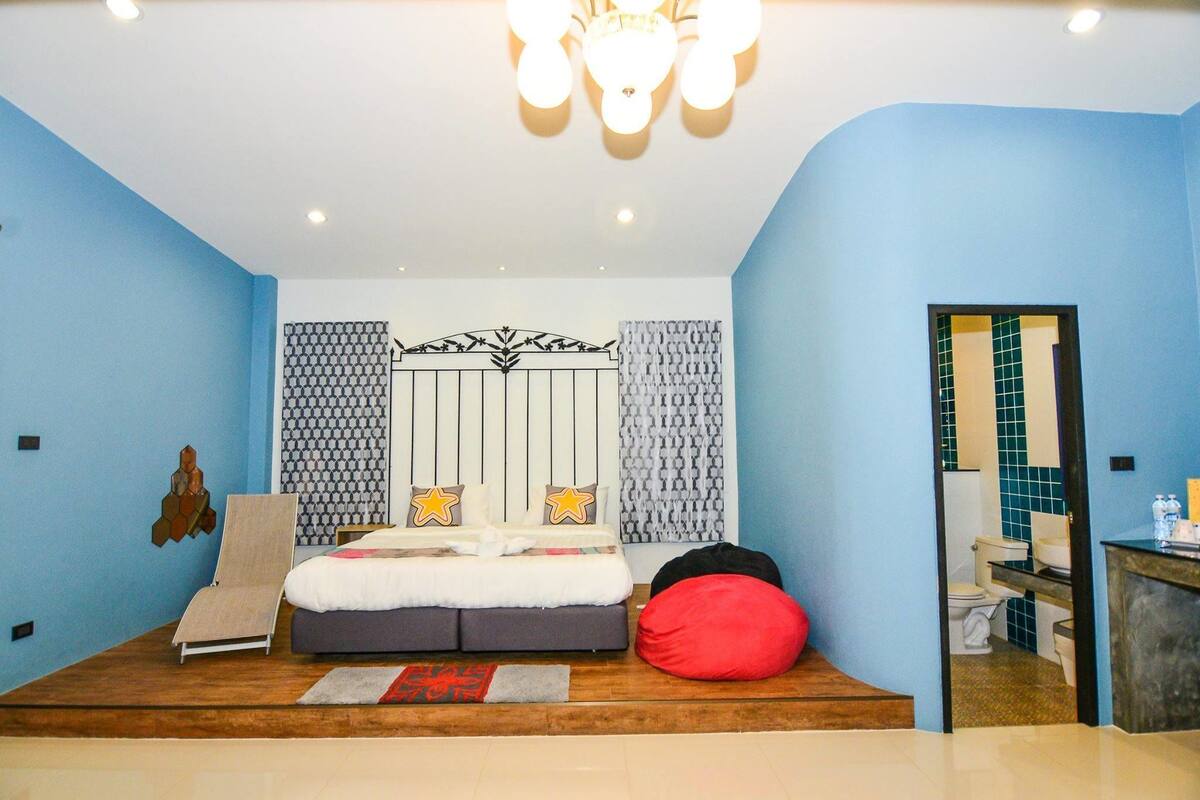 A bedroom with a bed, a red bean bag, a wooden floor, and a bathroom visible through an open door.