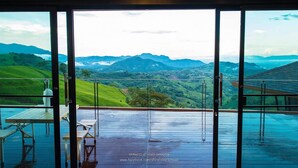 A room with a large sliding glass door leading to a balcony with a view of green hills and mountains. - Phu Sandao Resort (Khao Kho)