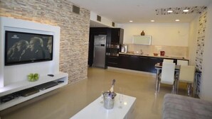 A modern living room with a flat-screen TV mounted on the wall, a white coffee table in the center, and a kitchen in the background with dark cabinetry and a white countertop. - La Palmora Suites (Eilat)