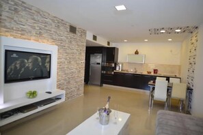 A modern living room with a flat-screen TV mounted on the wall, a white coffee table in the center, and a kitchen in the background with dark cabinetry and a white countertop. - La Palmora Suites (Eilat)