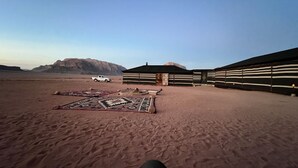 A desert landscape with a white pickup truck, a tent-like structure, and a patterned rug on the sand.