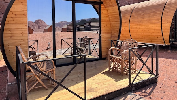 A circular wooden structure with a glass door, furnished with wicker chairs and a small table, set against a desert landscape with rock formations.