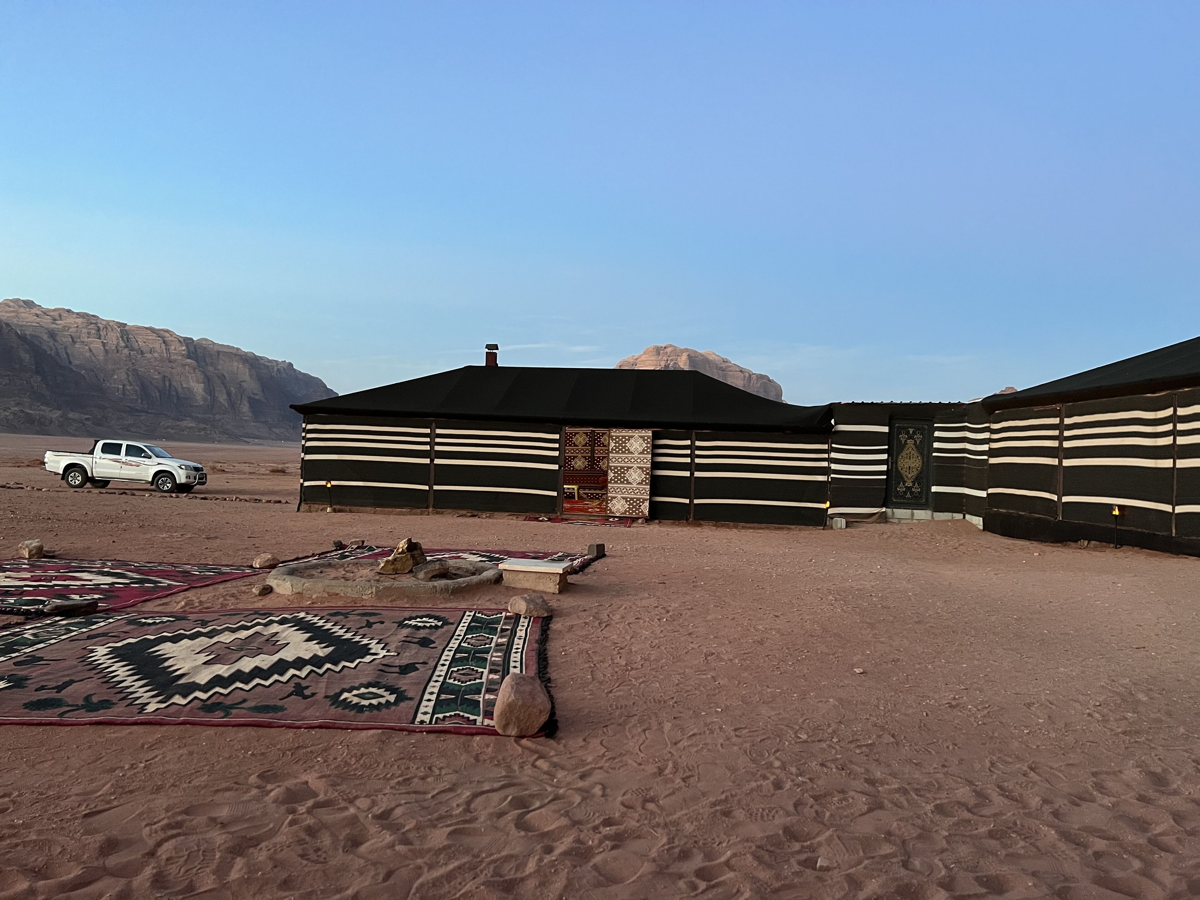 A desert setting with a building featuring horizontal wooden slats and a black roof. A white pickup truck is parked in front.