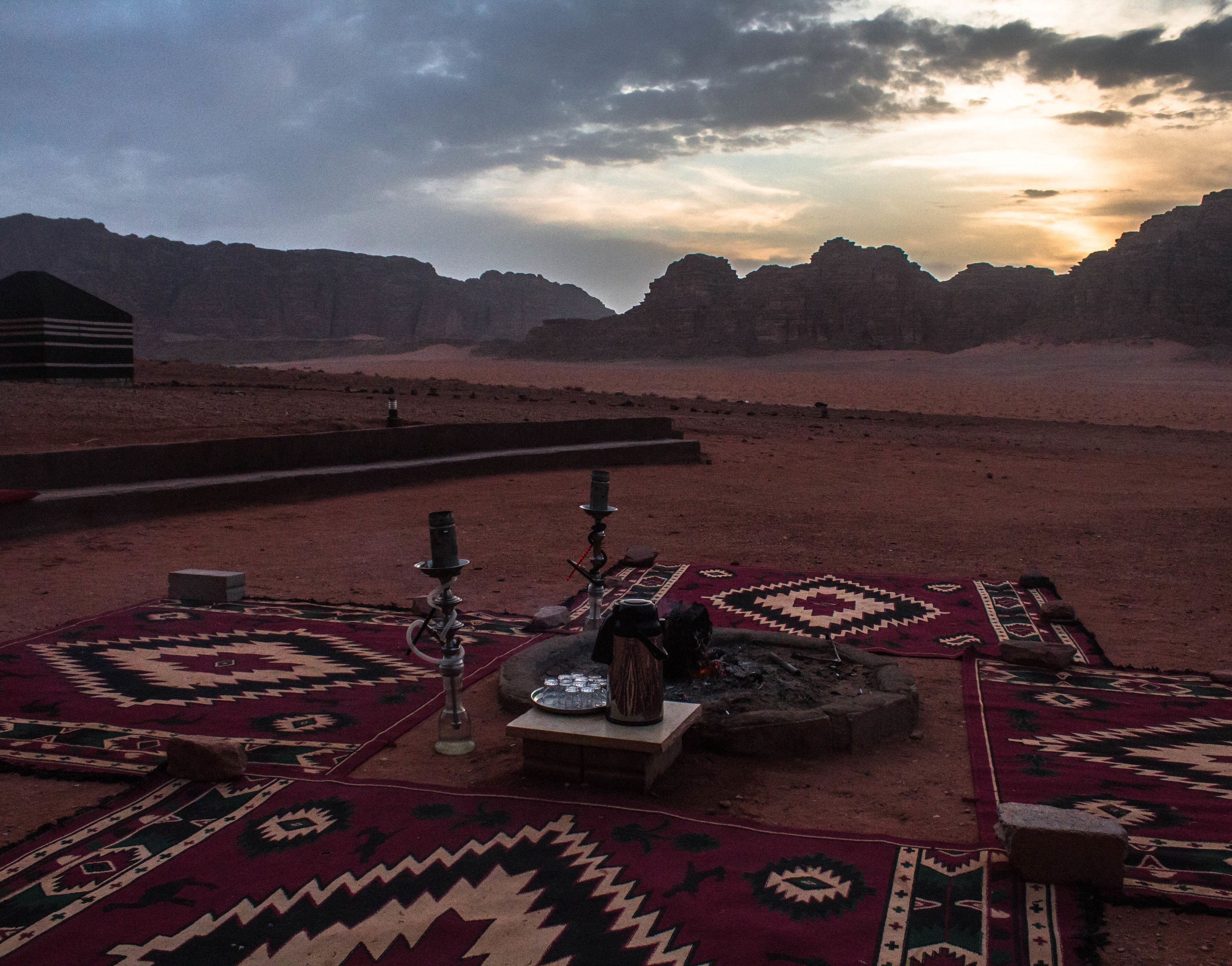 A desert landscape with a traditional setting featuring a patterned carpet, a hookah, and a small table.
