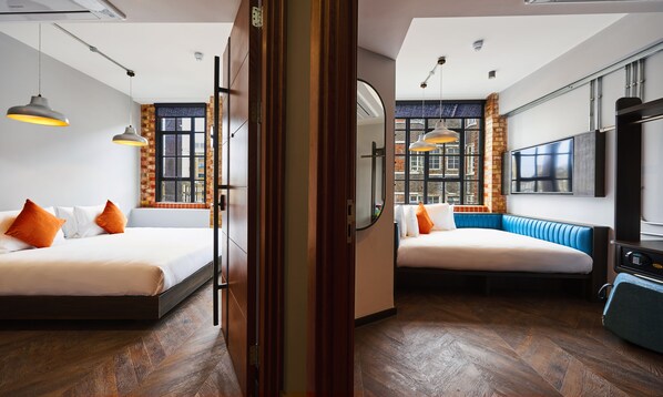 Habitación de hotel con dos camas, un banco azul, un televisor y un amplio ventanal con vista a edificios.