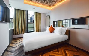 A modern hotel room with a large bed, a flat-screen TV, a large window with curtains, and a herringbone-patterned floor. - New Road Hotel (London)