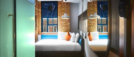 Una habitación de hotel moderna con una pared de ladrillo decorativa, una cama grande con ropa de cama blanca, una vista a la ventana azul y un espejo que refleja la habitación.