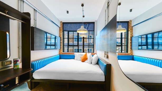 A modern hotel room with a large bed, a desk, and a view of the city through large windows. - New Road Hotel (London)