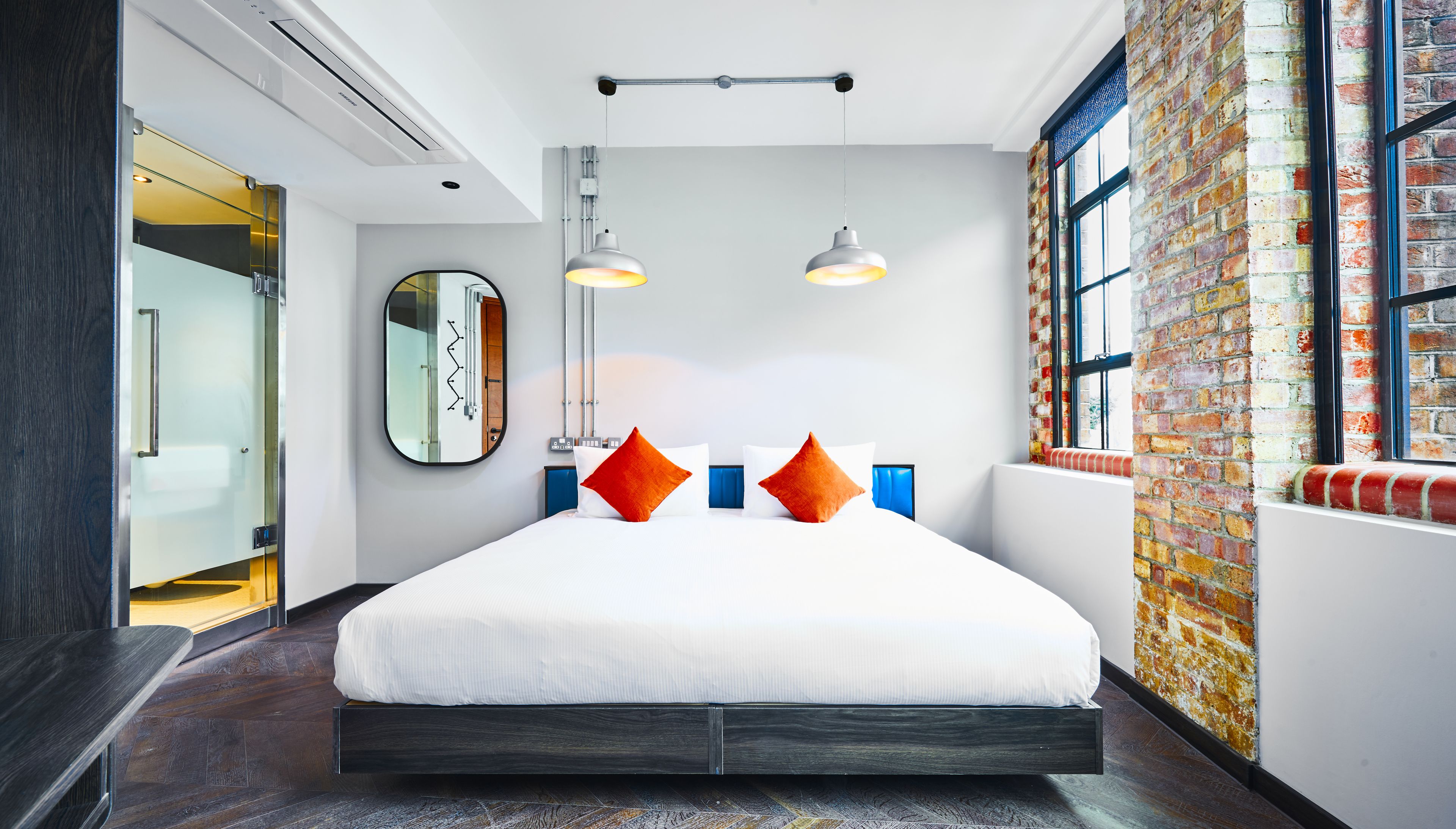 Une chambre d’hôtel moderne avec un grand lit, deux lampes suspendues, un miroir, un mur en briques et une fenêtre.