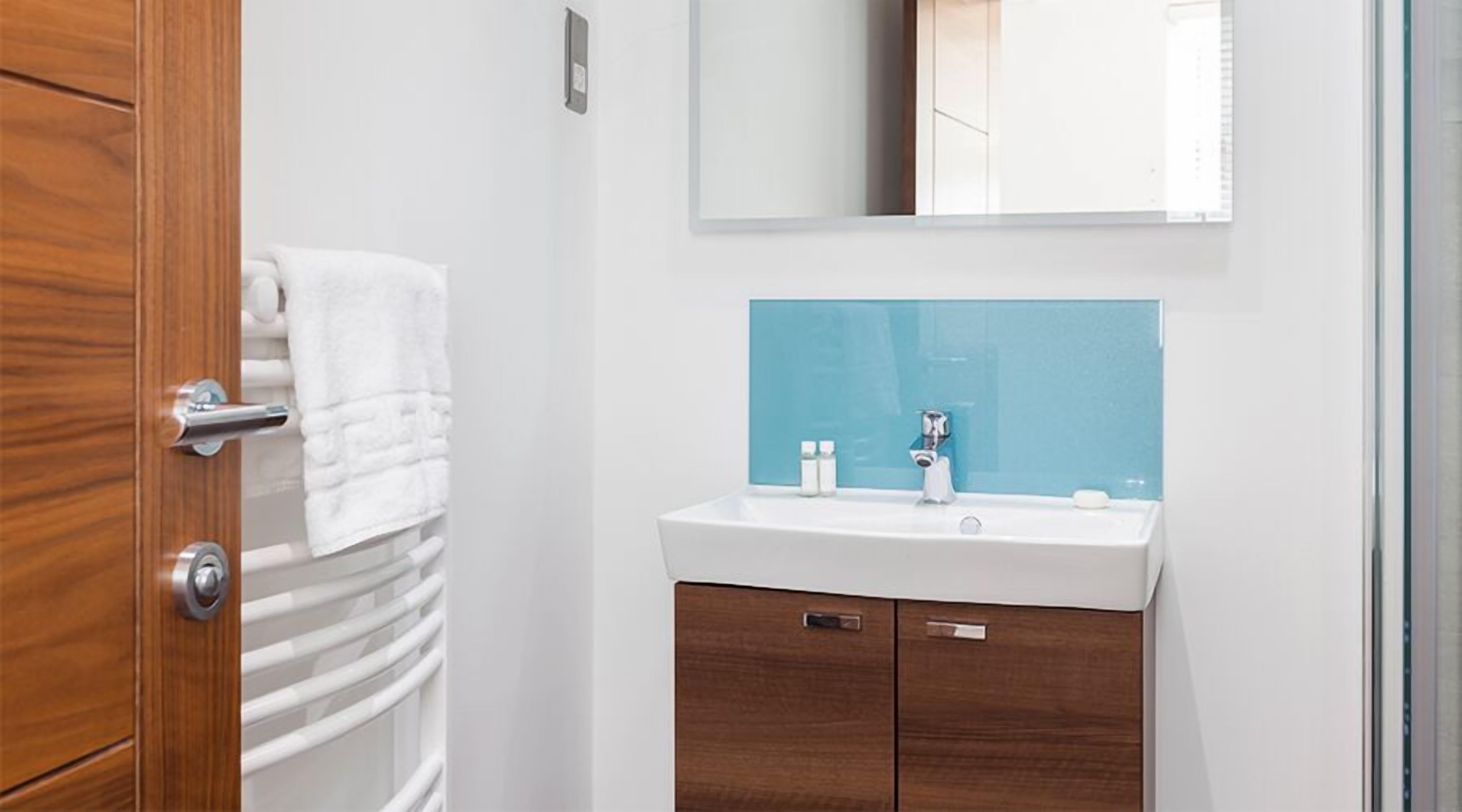A bathroom with a wooden door, a white towel hanging on the door, a white sink with a blue backsplash, and a mirror above the sink.