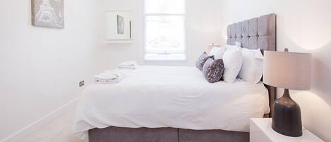 A bedroom with a bed, pillows, a nightstand, a lamp, and a window.