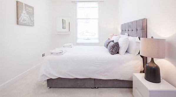 Une chambre à coucher avec un lit, des oreillers, une table de chevet, une lampe et une fenêtre.