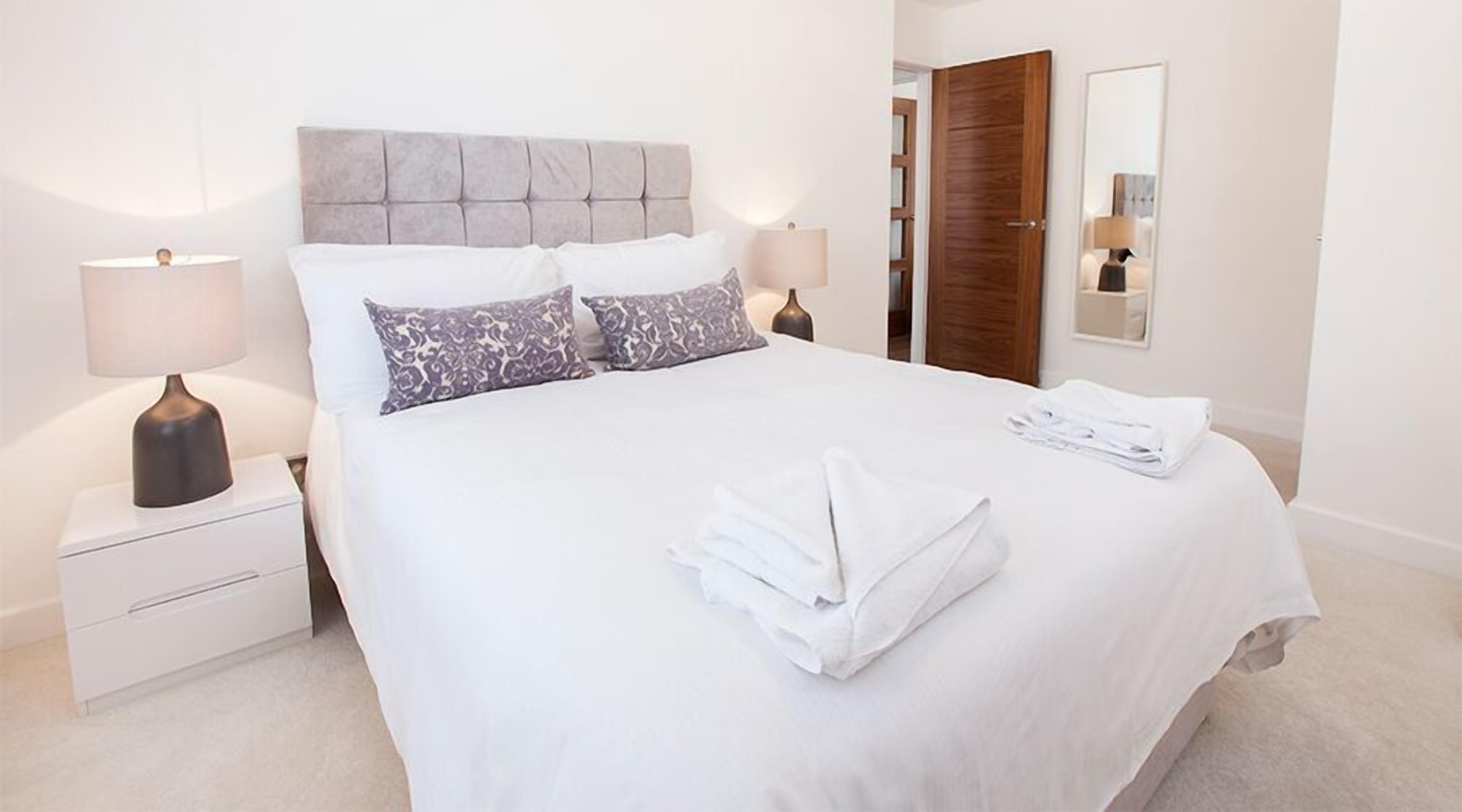 Una cama bien tendida con sábanas blancas y dos almohadas estampadas, una mesita de noche con una lámpara, y una puerta de madera al fondo.