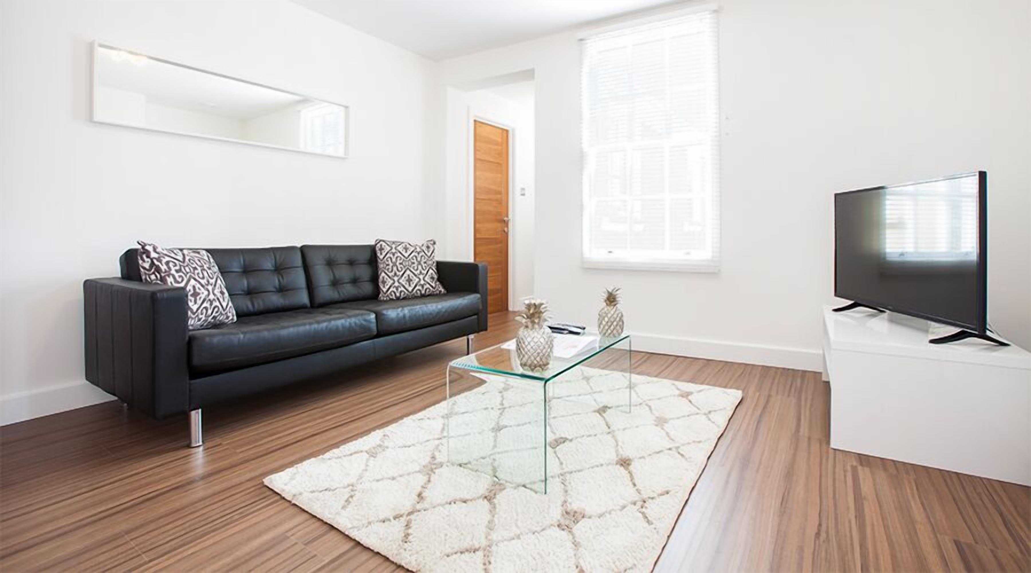 A modern living room with a black leather sofa, a glass coffee table, a flat-screen TV on a white stand, and a large window with blinds.