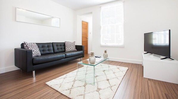 Un salon moderne avec un canapé en cuir noir, une table basse en verre, un téléviseur à écran plat posé sur un meuble blanc et une grande fenêtre équipée de stores.