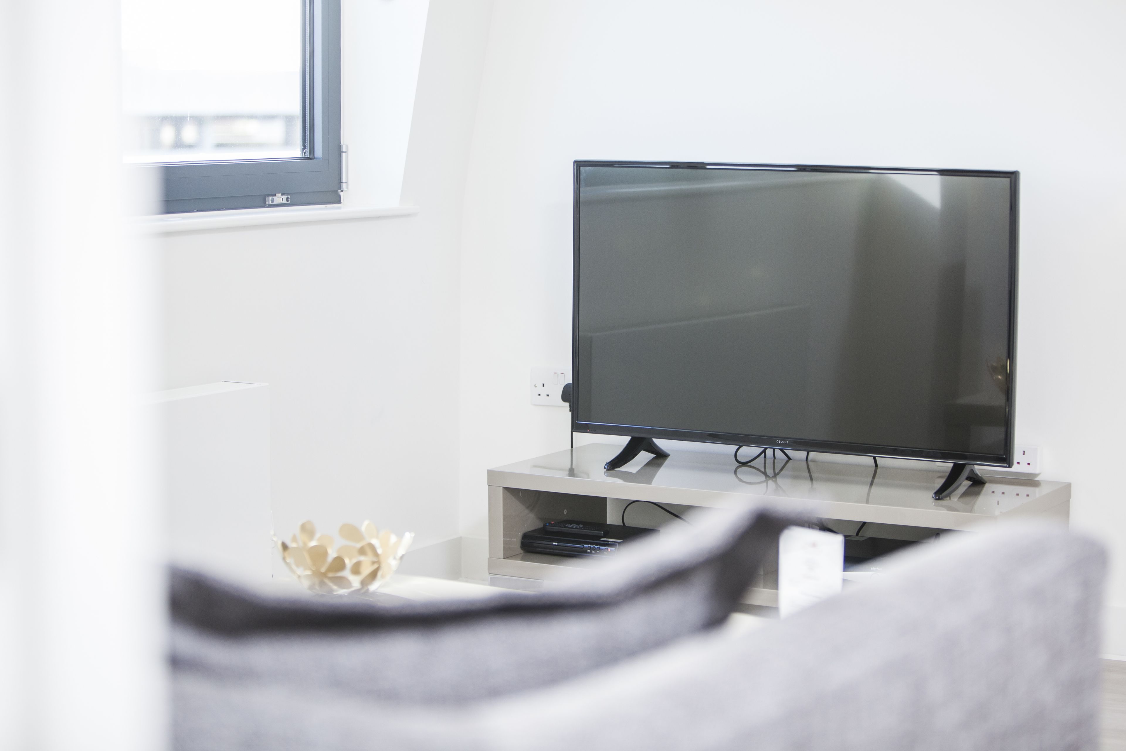 Una sala de estar moderna con una televisión de pantalla plana sobre un mueble bajo, una mesa de centro con un control remoto y una ventana con persianas.
