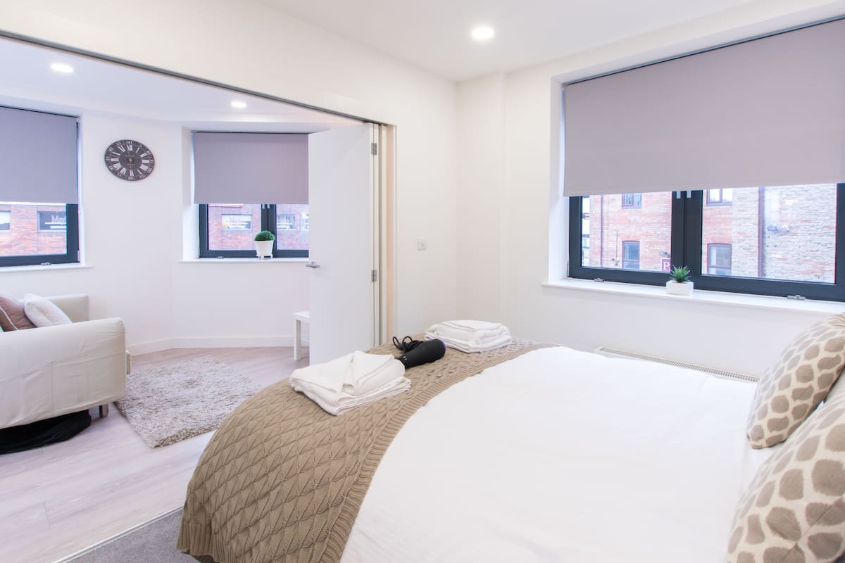 Quarto de hotel moderno com uma cama, um sofá, uma janela grande com vista para um edifício de tijolos e um relógio na parede.