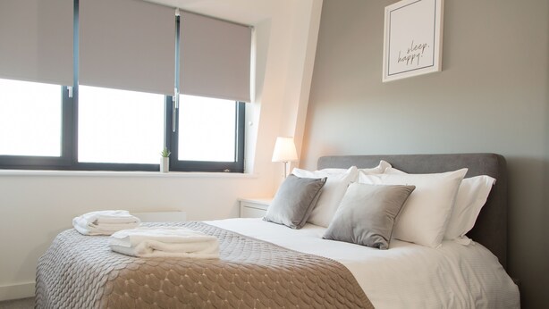 A neatly made bed with a beige quilt, a grey headboard, and a bedside lamp.