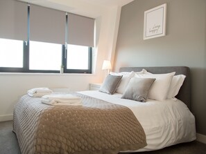 A neatly made bed with a beige quilt, a grey headboard, and a bedside lamp. - City Stay Apartments - Platform (Bedford)