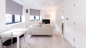 A modern, well-lit room with a white sofa, a dining table with chairs, and a large window with blinds.