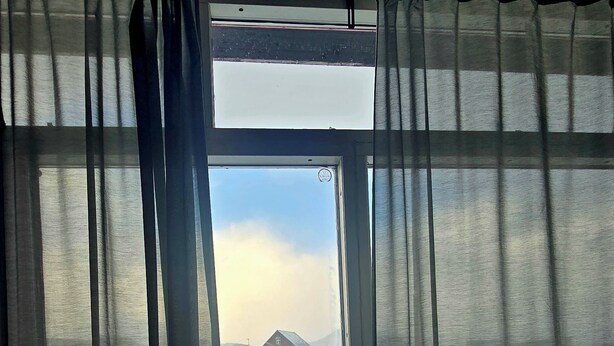 A window with curtains partially drawn, revealing a snowy outdoor scene with buildings and a clear sky. - Reykjanes Guesthouse (Grindavik)