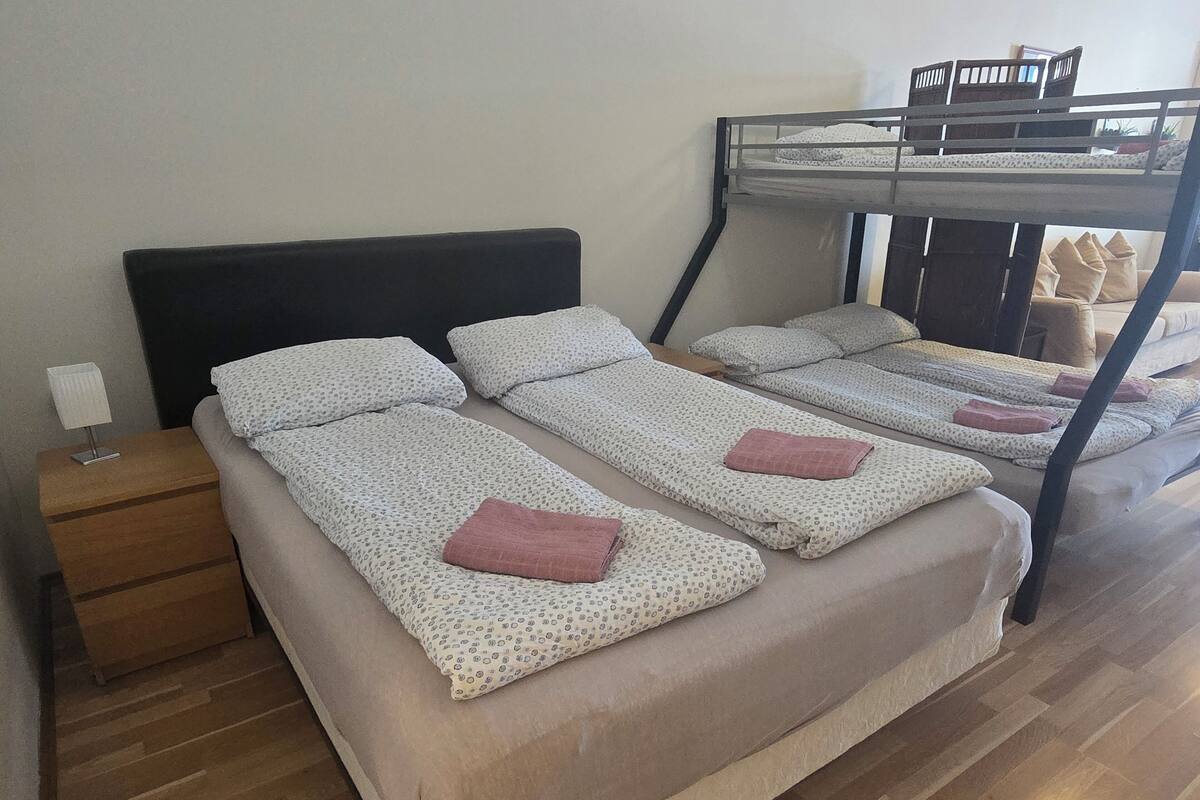 A room with a bunk bed, two single beds, a wooden nightstand, and a lamp.