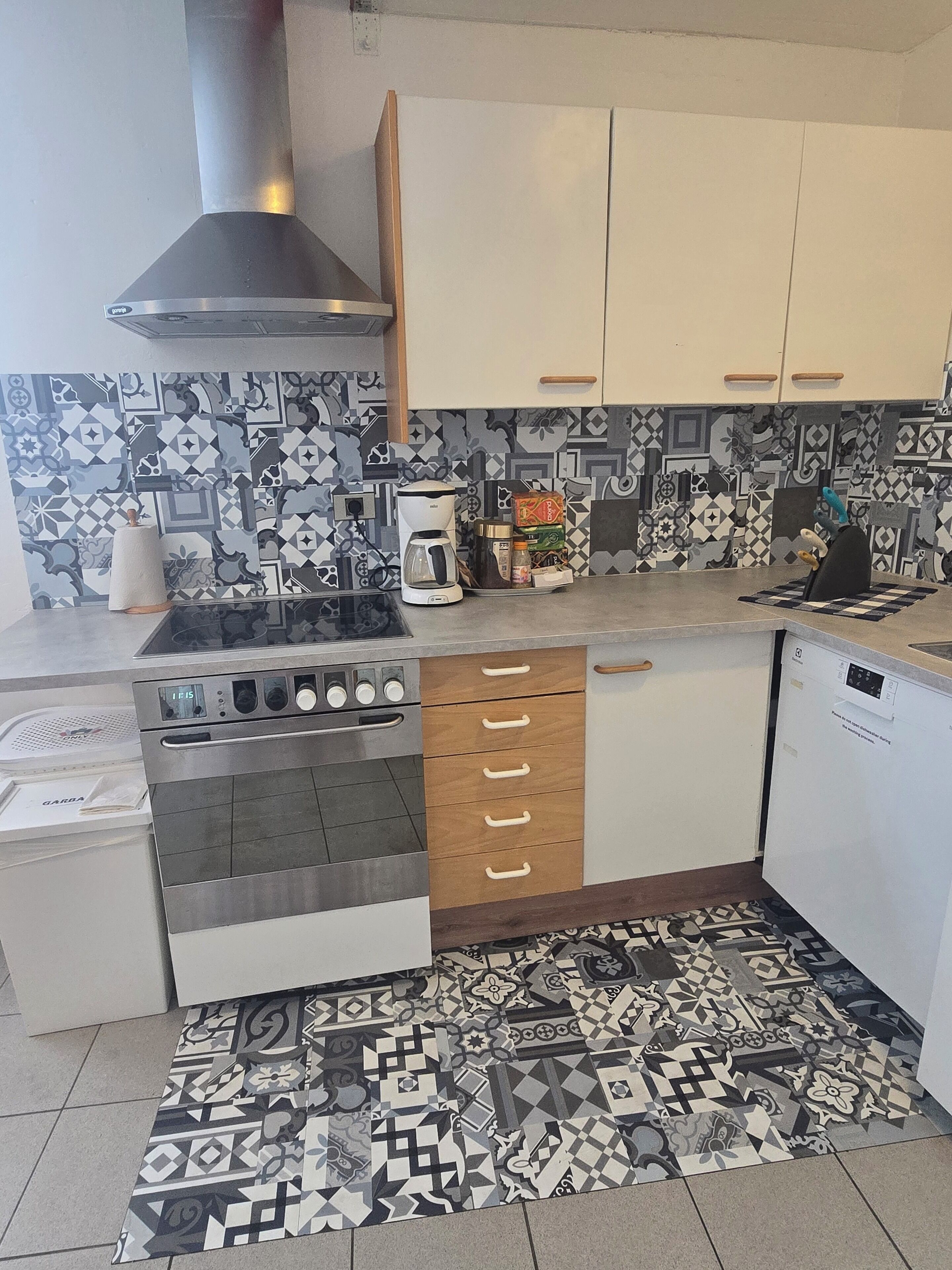 A compact kitchen with a gas stove, oven, and coffee maker. It features patterned tiles on the backsplash and a patterned rug on the floor.