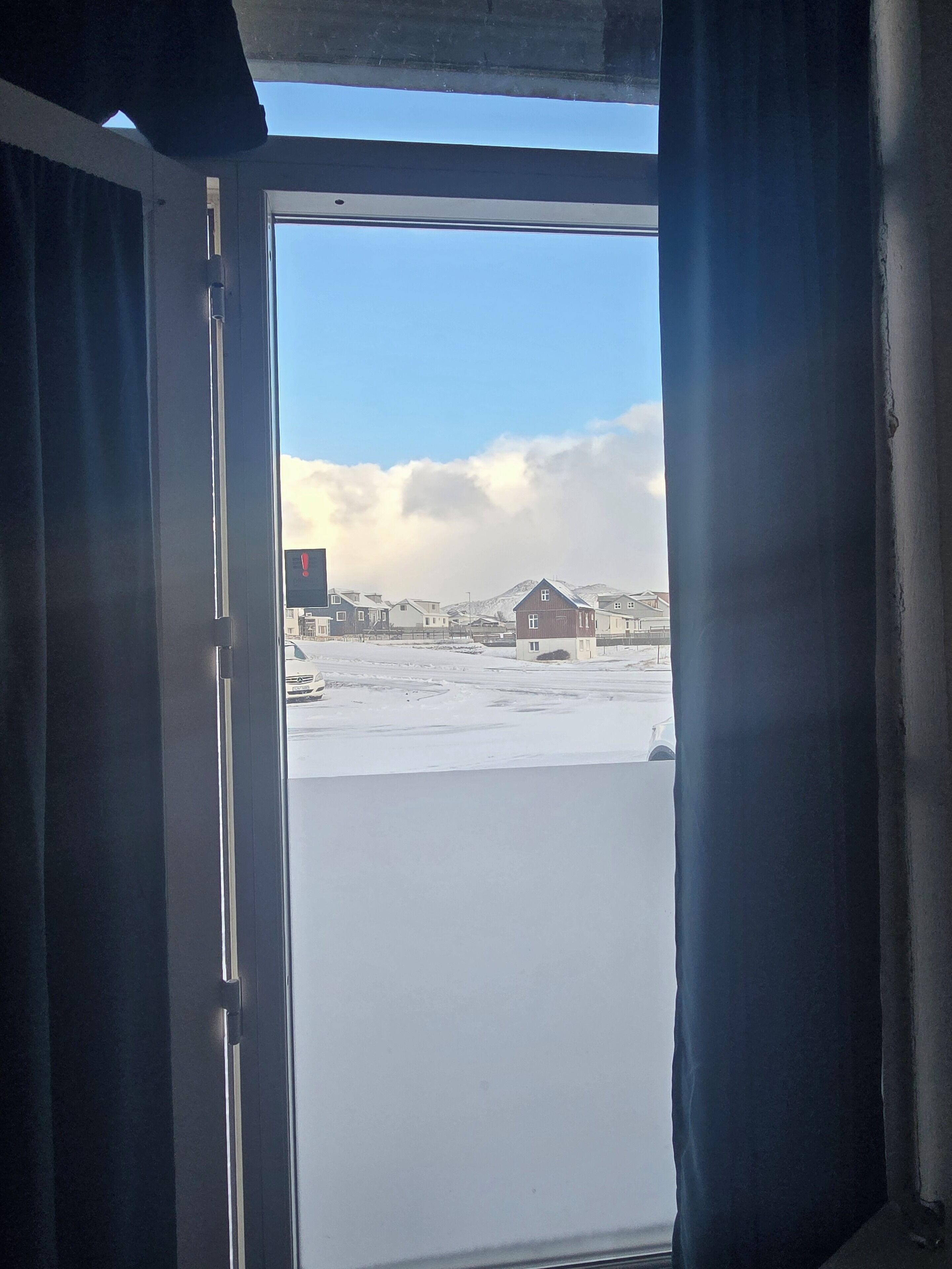A view through a window with curtains, revealing a snowy landscape with buildings and a clear sky.