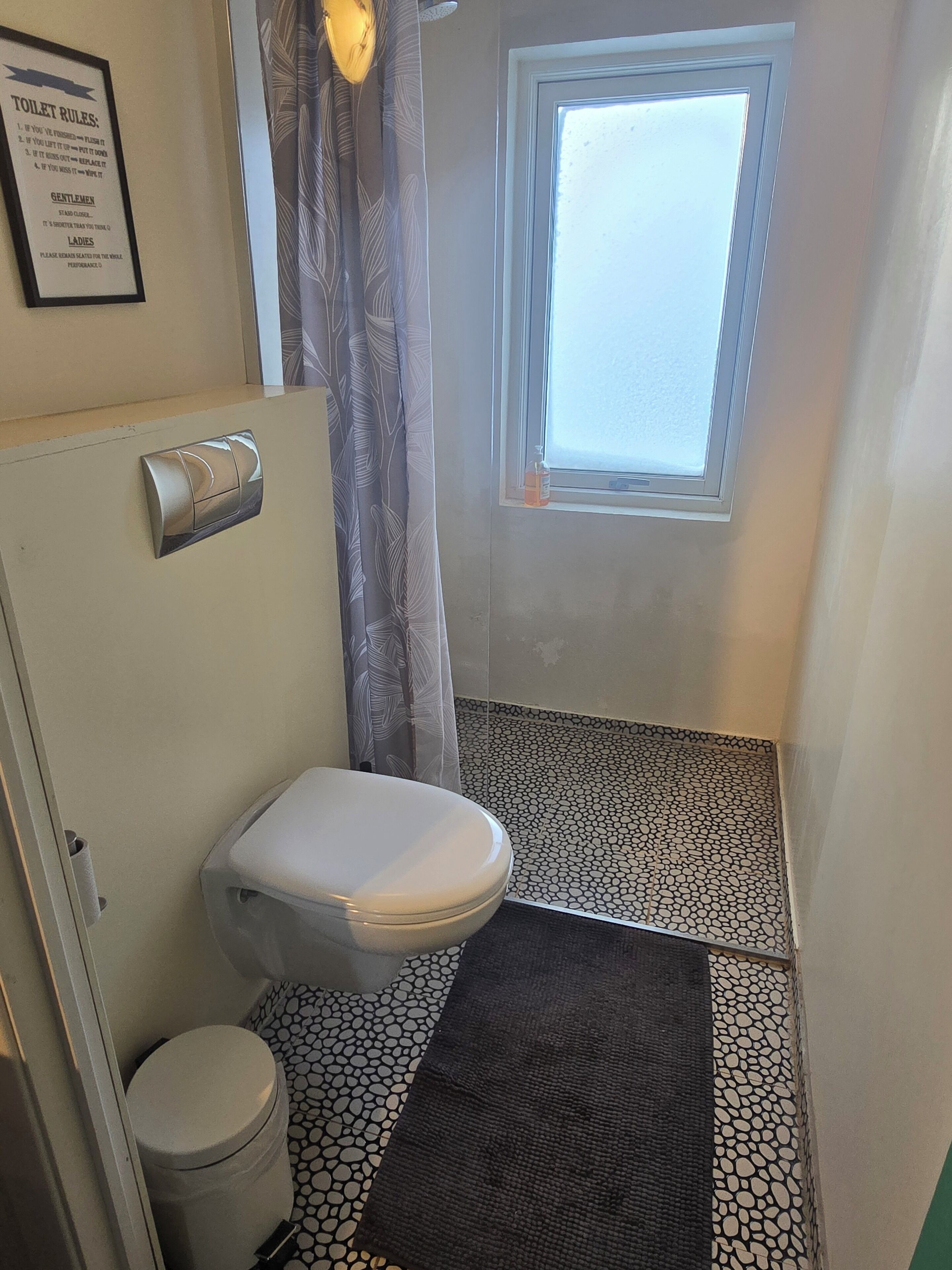 Ein Badezimmer mit Toilette, einem Händetrockner, einem Fenster und einem gefliesten Boden.
