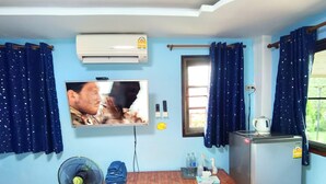 Ein Zimmer mit Fernseher an der Wand, einem kleinen Holztisch, einem Kühlschrank, einem Ventilator und Vorhängen.