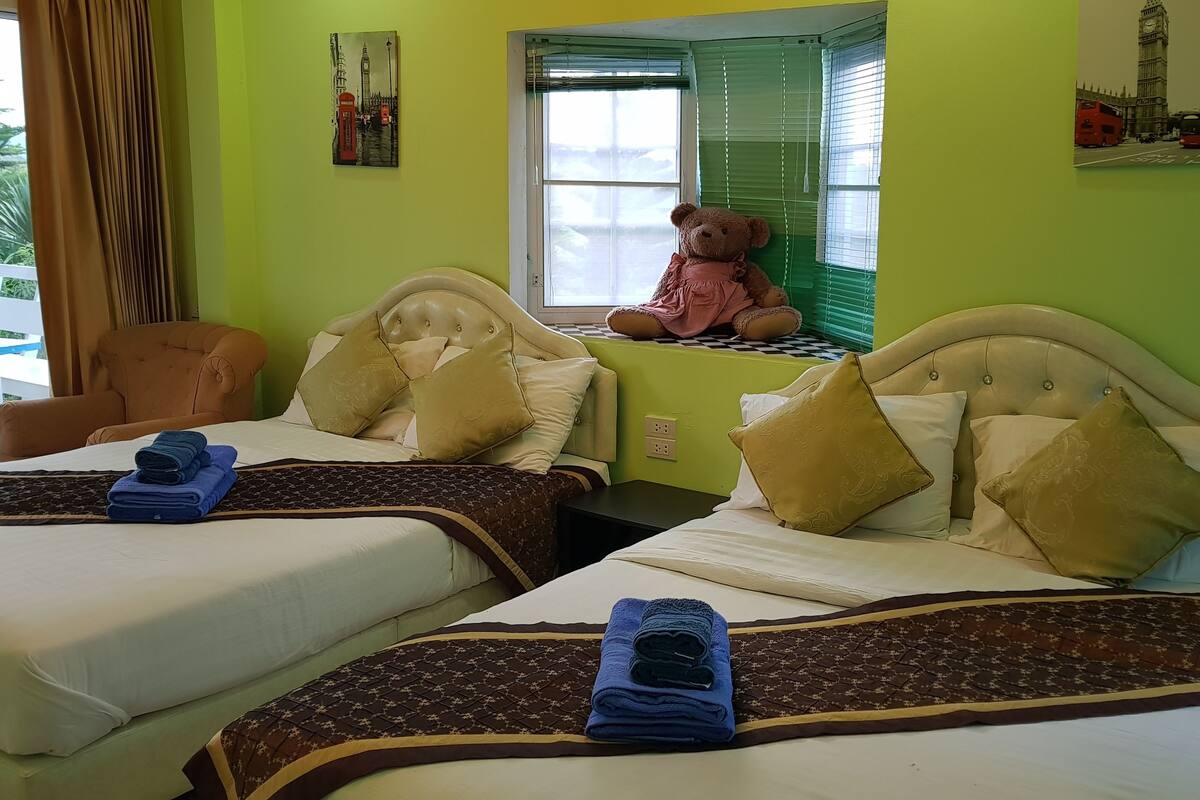 Ein Hotelzimmer mit zwei Betten, jeweils mit Kopfteil, einem Fenster mit Jalousien, einem Teddybären auf dem Fensterbrett und einem Bild eines roten Telefonhäuschens.