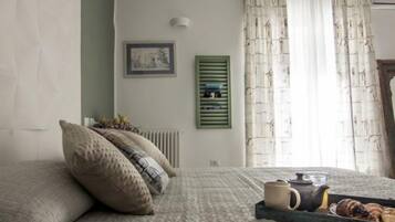 Une chambre à coucher avec un lit, des oreillers, un radiateur, un tableau au mur, une fenêtre avec des rideaux et un plateau avec une théière et des pâtisseries.