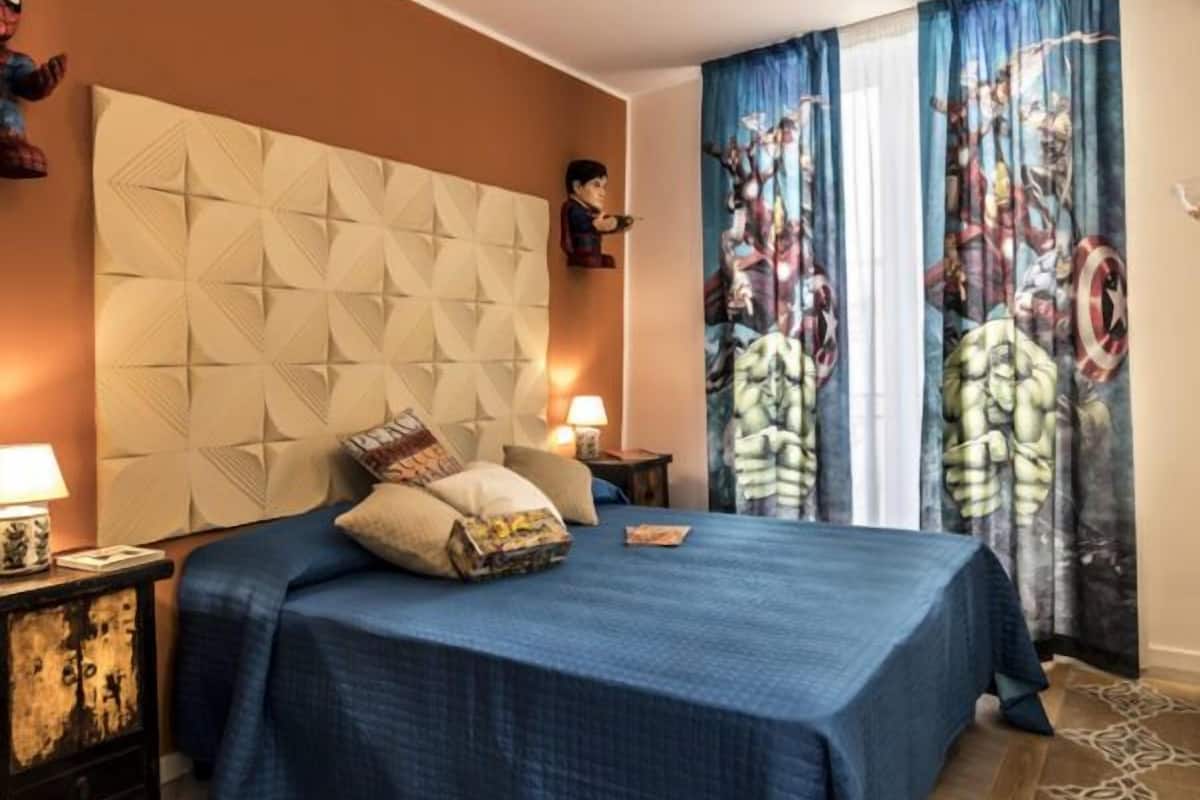 A bedroom with a large bed, a headboard, a nightstand, a lamp, and curtains with a superhero design.